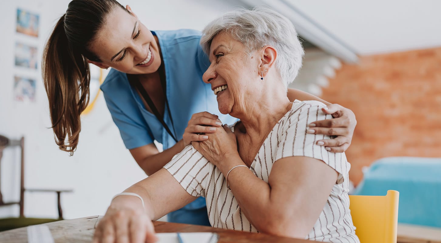 A nurse assists an elderly woman at a table, providing care and support in a healthcare setting.